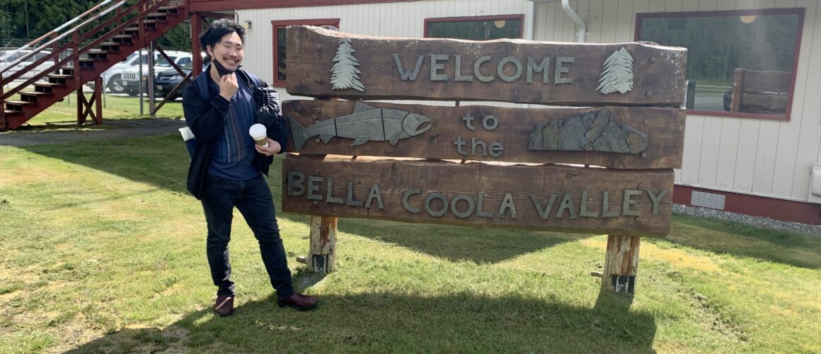 Welcome sign to Bella Coola Valley at airport, Ben Chung stands in front of sign